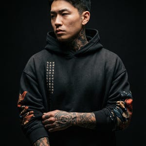 Pu&ograve; includere: Una felpa con cappuccio nera con testo giapponese sul davanti e opere d'arte dettagliate sulle maniche. Le maniche presentano un design con accenti rossi, arancioni e blu. La felpa con cappuccio &egrave; indossata da una persona con tatuaggi sul collo e sulle braccia.