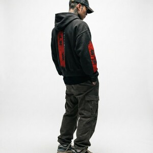 May include: A person wearing a black hoodie with red Japanese text on the back and sleeve. The person is also wearing a camouflage baseball cap, dark green cargo trousers, and grey and beige trainers.