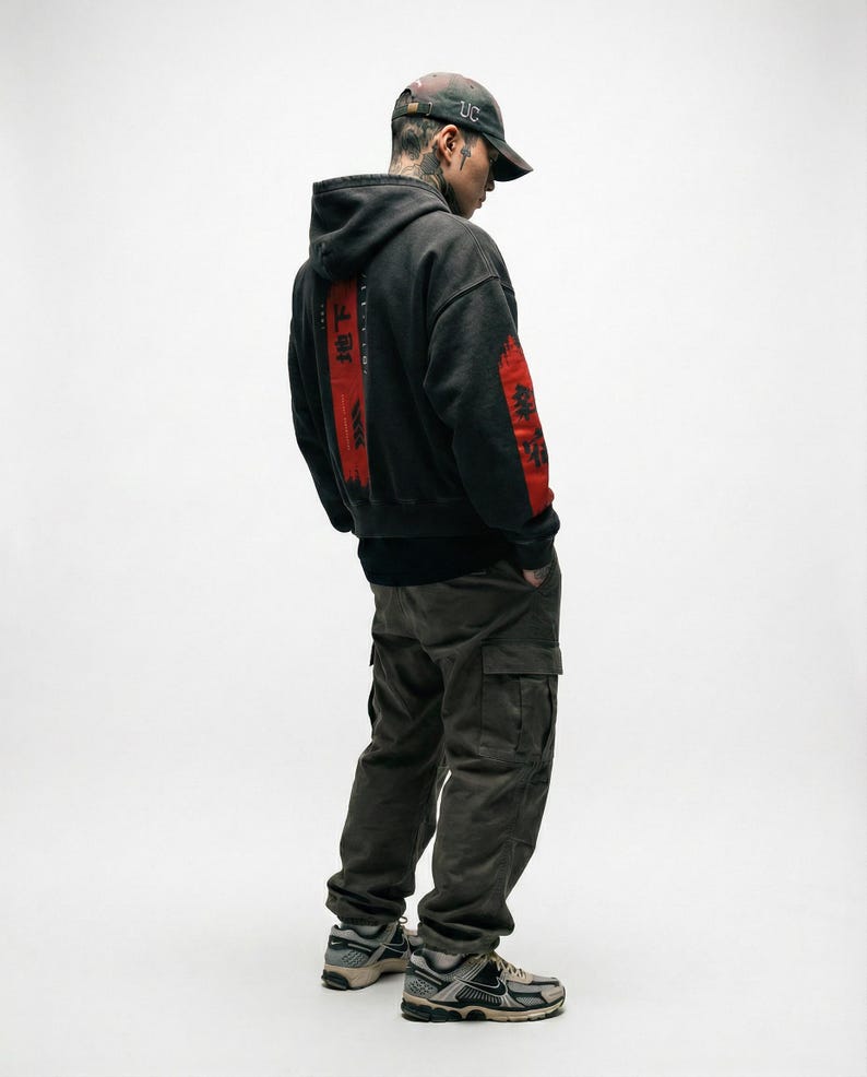 May include: A person wearing a black hoodie with red Japanese text on the back and sleeve. The person is also wearing a camouflage baseball cap, dark green cargo trousers, and grey and beige trainers.