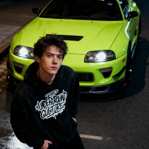 May include: A black hooded sweatshirt featuring the white text Underground Culture in a stylized font on the chest, worn with black pants, positioned in front of a bright lime green sports car with a rear wing and a windshield banner.