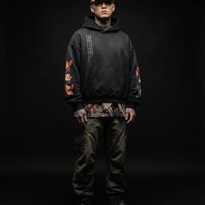 Pu&ograve; includere: Una persona indossa una felpa con cappuccio nera con dettagli floreali e testo giapponese, abbinata a pantaloni cargo verde oliva e un berretto da baseball. La felpa ha una tasca frontale e viene indossata sopra una camicia fantasia. La persona indossa anche scarpe da ginnastica nere.