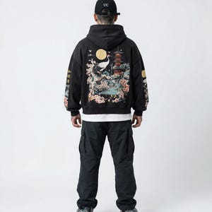 May include: Black hoodie with a Japanese-inspired design on the back, featuring a crane, pagoda, and cherry blossoms. The sleeves have additional Japanese text. The person is wearing black cargo pants and a black baseball cap.