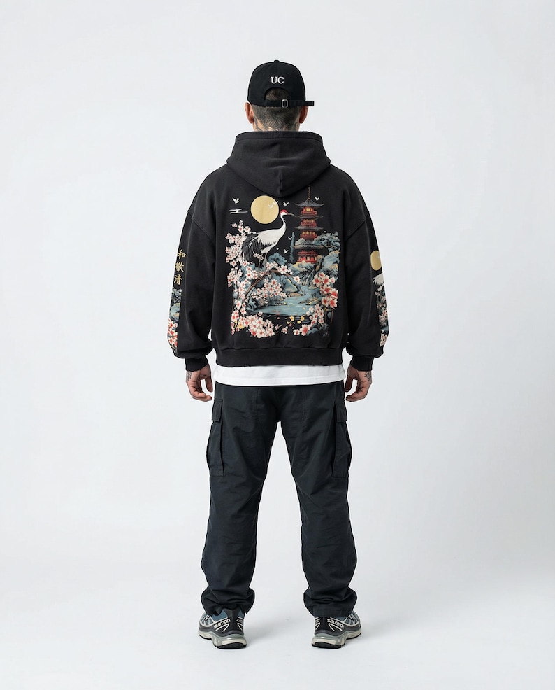 May include: Black hoodie with a Japanese-inspired design on the back, featuring a crane, pagoda, and cherry blossoms. The sleeves have additional Japanese text. The person is wearing black cargo pants and a black baseball cap.