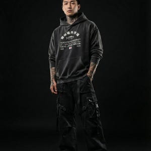 May include: A charcoal gray hooded sweatshirt featuring white text that reads Tsukiji Outer Market Tokyo Japan 2018 with a fish graphic, paired with black cargo pants that have multiple pockets, straps, and buckles on the legs and thighs.