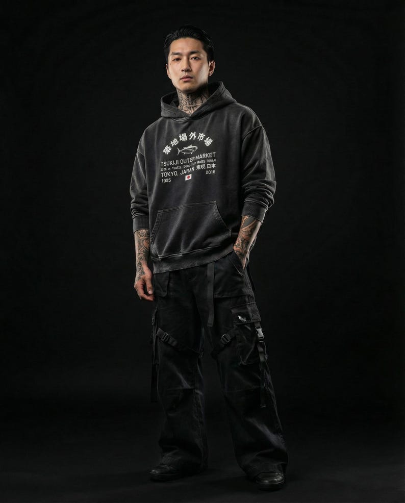 May include: A charcoal gray hooded sweatshirt featuring white text that reads Tsukiji Outer Market Tokyo Japan 2018 with a fish graphic, paired with black cargo pants that have multiple pockets, straps, and buckles on the legs and thighs.