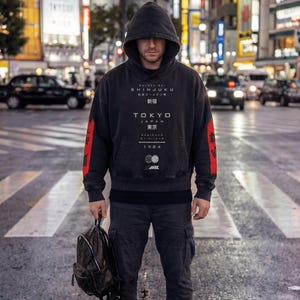 May include: A person in a black hoodie with red sleeves and "TOKYO JAPAN" text stands on a pedestrian crossing at night. They hold a backpack, wearing black trousers and trainers. The background shows illuminated city buildings.