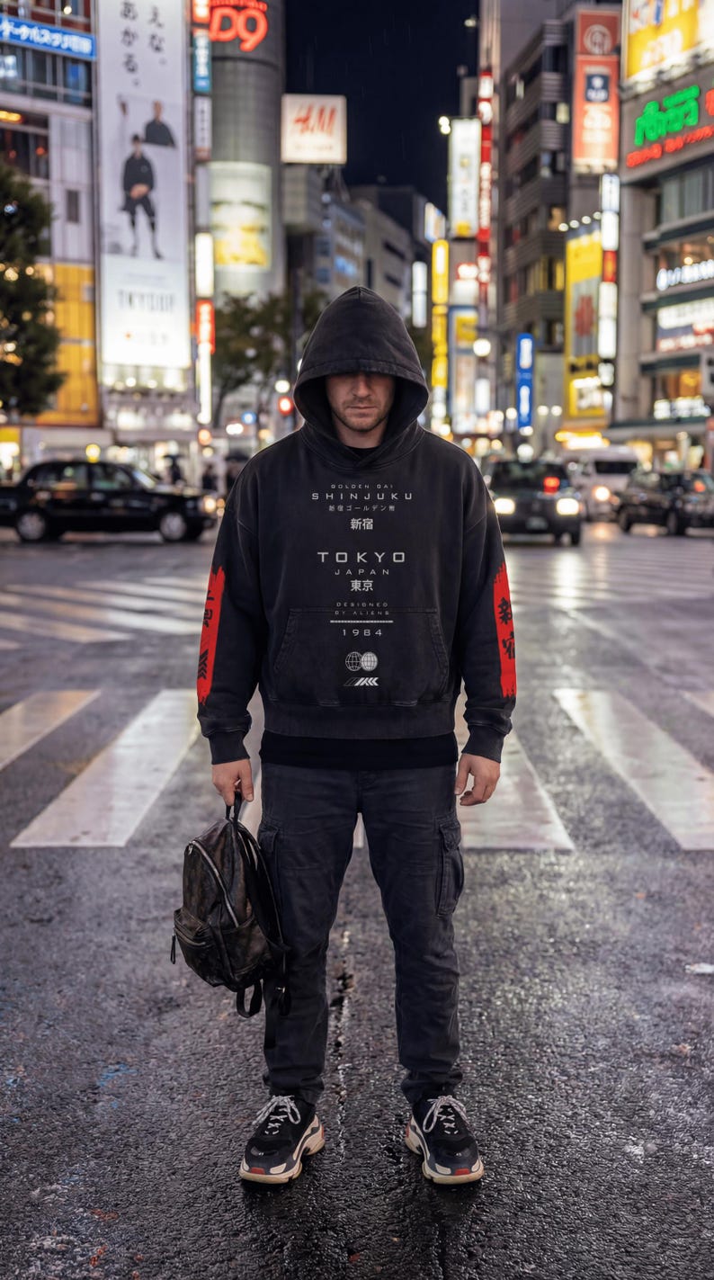 May include: A person in a black hoodie with red sleeves and "TOKYO JAPAN" text stands on a pedestrian crossing at night. They hold a backpack, wearing black trousers and trainers. The background shows illuminated city buildings.