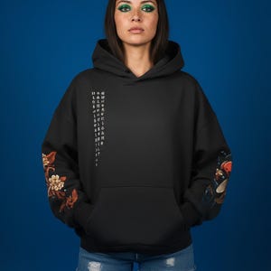 Pu&ograve; includere: Felpa con cappuccio nera con tasca frontale e cappuccio con coulisse. Le maniche presentano disegni floreali e di pesci koi. Il testo giapponese &egrave; stampato verticalmente sul lato sinistro. La modella indossa pantaloncini di jeans blu.