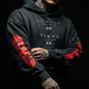 May include: Dark grey hoodie with distressed details and Japanese text. The hoodie features red accents on the sleeves with black graphic designs. The text on the front reads "SHINJUKU" and "TOKYO".
