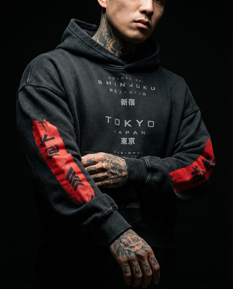 May include: Dark grey hoodie with distressed details and Japanese text. The hoodie features red accents on the sleeves with black graphic designs. The text on the front reads "SHINJUKU" and "TOKYO".