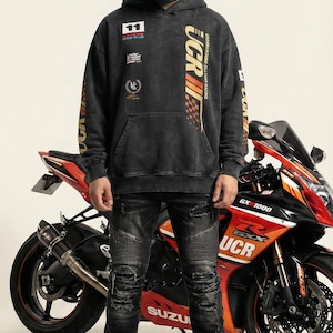 May include: A person wearing a black hoodie with "UCR" and racing graphics, distressed black jeans, and a motorcycle helmet. A red and black Suzuki GSX-1000 motorcycle is behind the person.