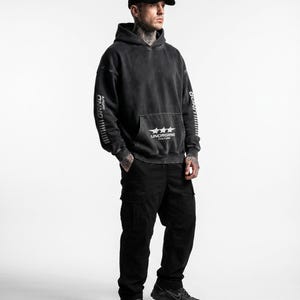 May include: A person wearing a black hoodie with the text "UNDERGRND CULTURE" and three stars on the front pocket. The hoodie has "GRND culture" printed on the sleeves. The person is also wearing black cargo pants and a black baseball cap.