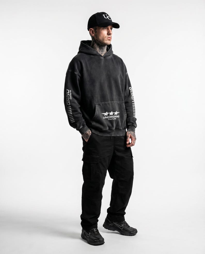 May include: A person wearing a black hoodie with the text "UNDERGRND CULTURE" and three stars on the front pocket. The hoodie has "GRND culture" printed on the sleeves. The person is also wearing black cargo pants and a black baseball cap.