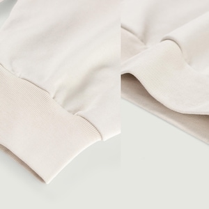 May include: Close-up of a cream-colored sweatshirt sleeve and cuff. The cuff is ribbed and the sleeve is made of a soft, smooth fabric. The background is a solid, light color.