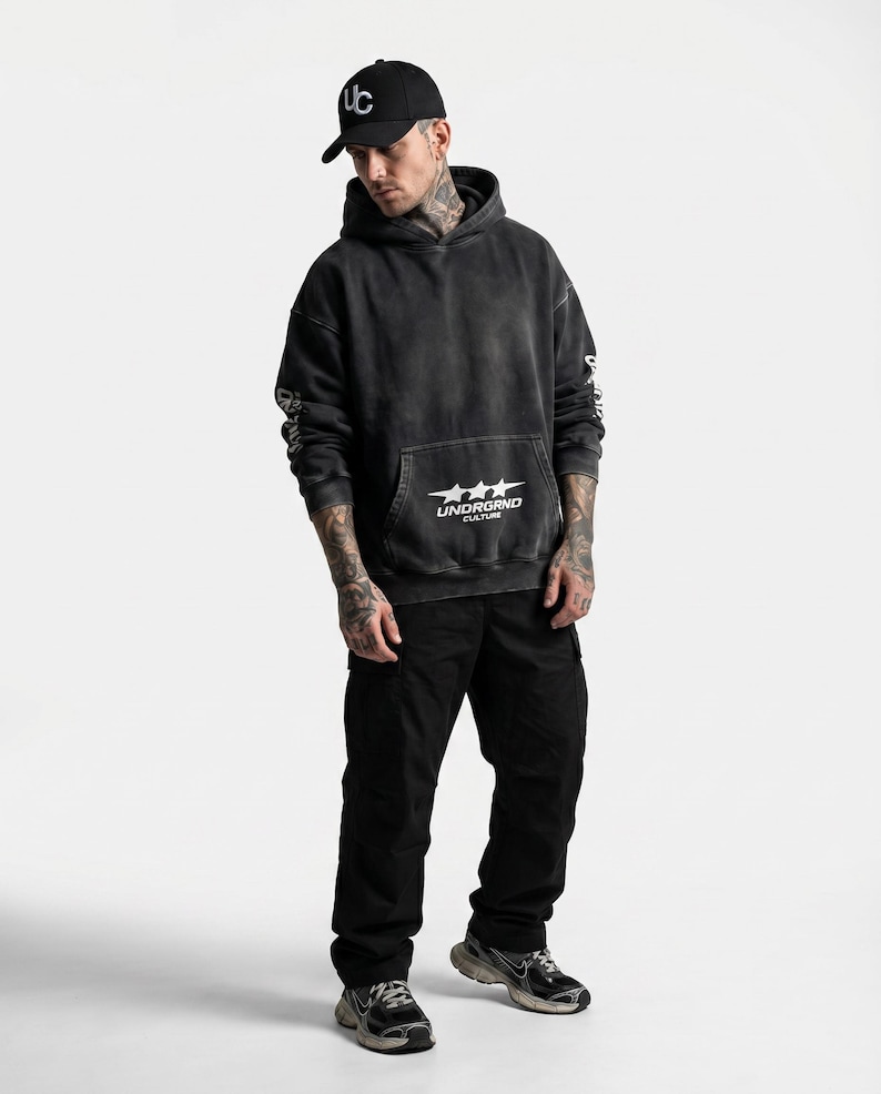 May include: A person wearing a black hoodie with the text "UNDRGRND CULTURE" and a black baseball cap with a white "UC" logo. The person is also wearing black cargo pants and black and gray sneakers.