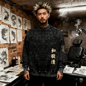 May include: A black and gray acid wash sweatshirt with gold Japanese characters. The sweatshirt has a crew neck and long sleeves. The background features tattoo art on the wall and a tattoo artist's workspace.