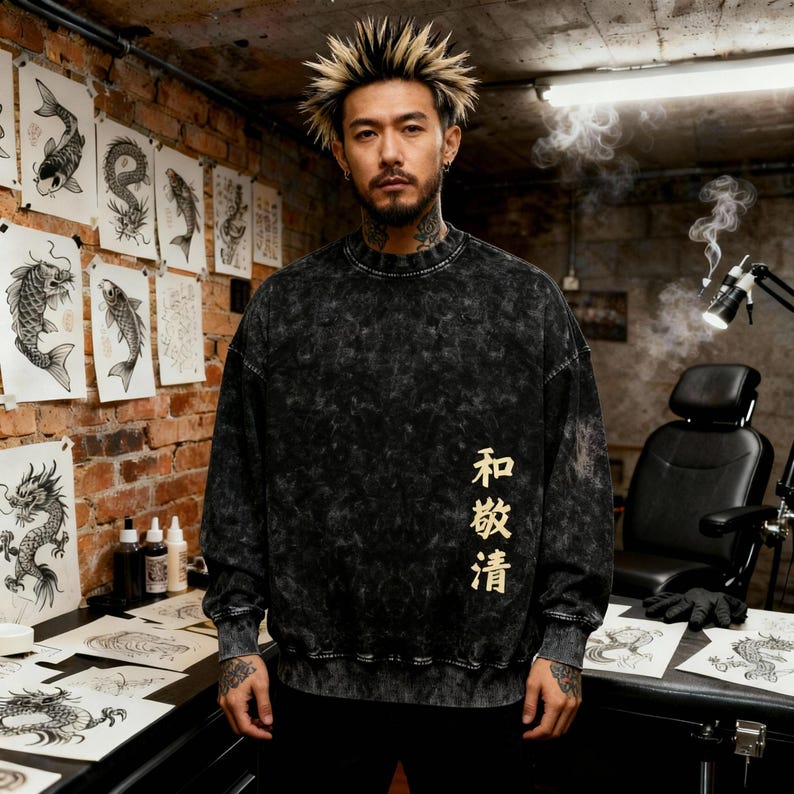 May include: A black and gray acid wash sweatshirt with gold Japanese characters. The sweatshirt has a crew neck and long sleeves. The background features tattoo art on the wall and a tattoo artist's workspace.