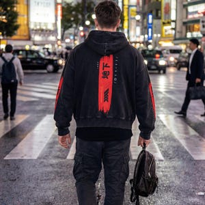 May include: A person crossing a street at night, wearing a black hoodie with red details and Japanese text. The person is also wearing black cargo trousers and trainers, carrying a black backpack.