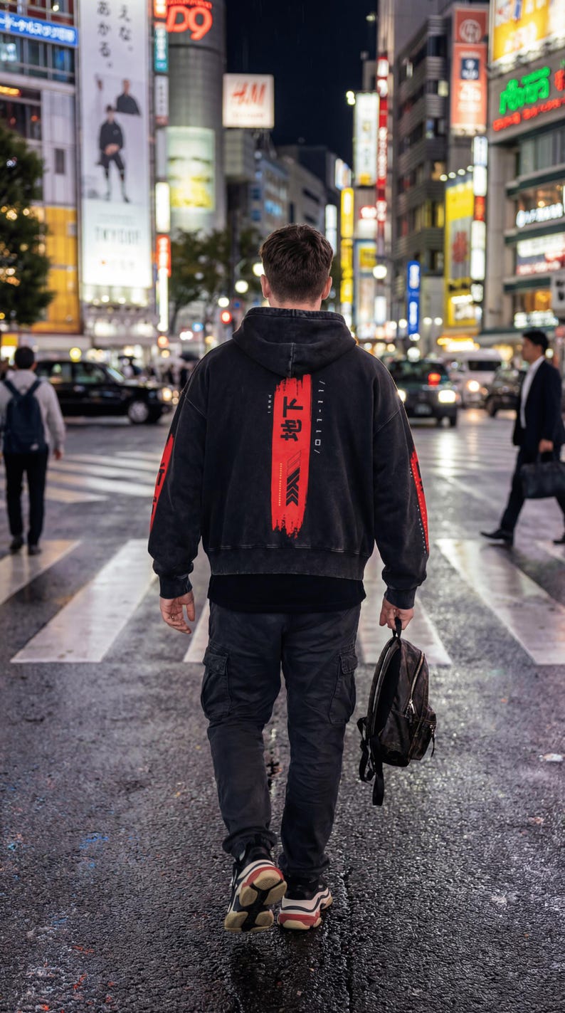 May include: A person crossing a street at night, wearing a black hoodie with red details and Japanese text. The person is also wearing black cargo trousers and trainers, carrying a black backpack.