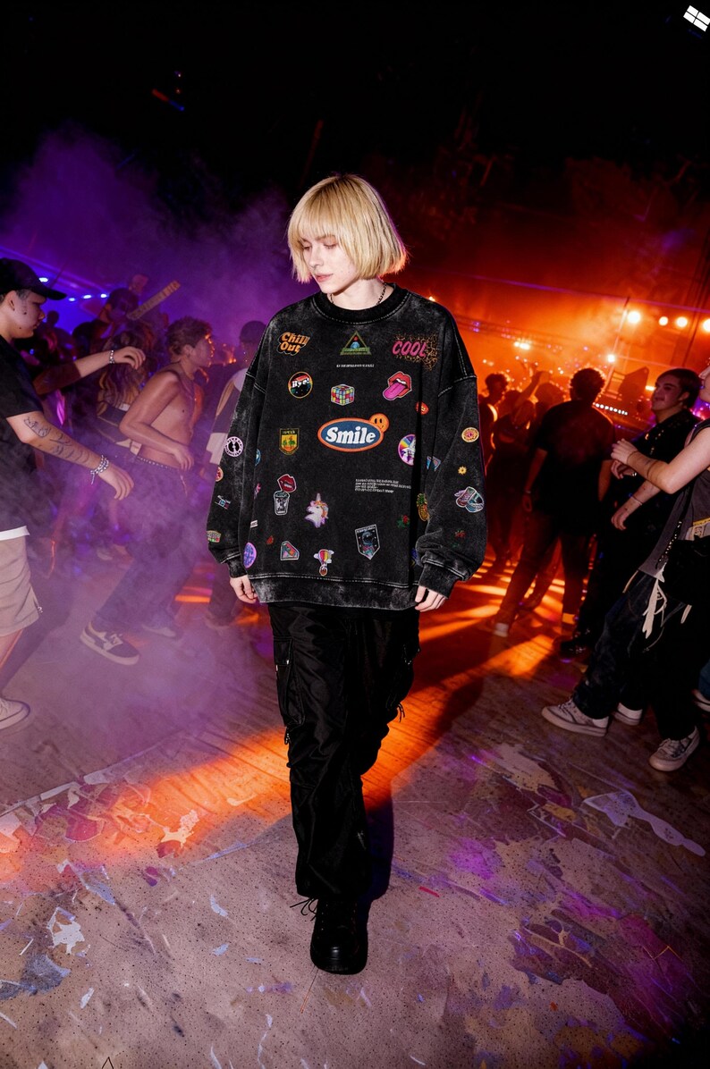 Pu&ograve; includere: Felpa nera oversize con varie toppe, tra cui la parola "Smile". La persona indossa pantaloni neri e scarpe nere. Lo sfondo mostra un evento affollato con persone e luci colorate.