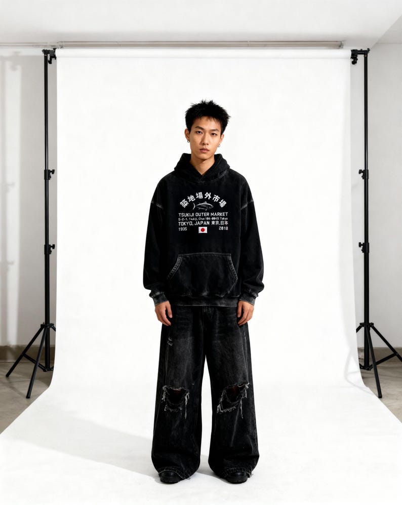 May include: A black acid-wash hooded sweatshirt featuring a white graphic print that reads Tsukiji Outer Market, Tokyo, Japan, with a fish illustration and a small Japanese flag. The hoodie is paired with matching black distressed wide-leg denim jeans.