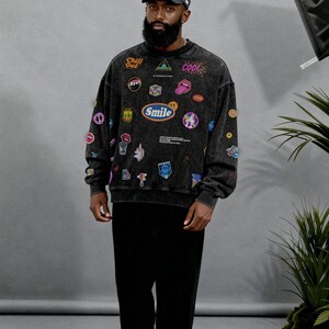 Pu&ograve; includere: Una felpa nera con diverse toppe colorate e le parole "Chill Out", "Cool" e "Smile". La persona indossa anche pantaloni neri, un berretto da baseball bianco e nero e scarpe da ginnastica grigie e bianche.