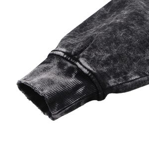 May include: Close-up of a black and gray acid wash sweatshirt sleeve. The cuff is ribbed and has a distressed, worn look. The fabric has a textured appearance with a faded, mottled pattern.