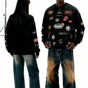 Pu&ograve; includere: Due persone indossano felpe nere con toppe colorate e le parole "Chill Out", "Cool" e "Smile". Indossano anche jeans blu con un design sfumato marrone. Lo sfondo &egrave; bianco.