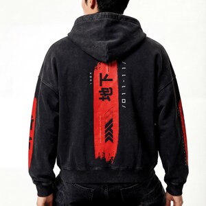 May include: Black hooded sweatshirt with red graphic elements and Japanese text on the back. The design includes vertical text and arrows. The sleeves also have red accents. The sweatshirt is made of a washed, textured fabric.