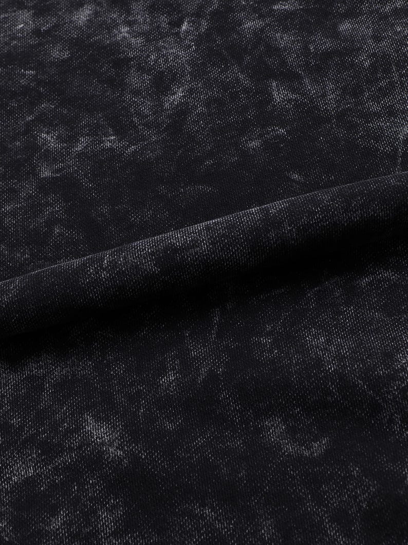 May include: Close-up of a black fabric with a textured, distressed appearance. The fabric has a mottled pattern with lighter gray areas, creating a worn or vintage aesthetic. The material is folded, showing the texture and drape of the fabric.