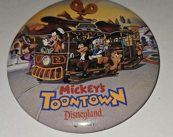 May include: A round button featuring Mickey Mouse and friends riding a trolley in a cartoon style. The trolley is red and brown with the words "Mickey's Toontown Disneyland" printed on the bottom. The button is a light gray color with a white border.