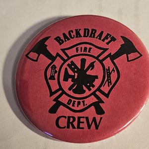 Peut inclure: Un bouton rond rouge avec l'inscription noire "BACKDRAFT FIRE DEPT. CREW". Le bouton présente un emblème de pompier noir avec des haches croisées et d'autres outils.