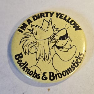 Op de afbeelding: Een ronde, gele button met zwarte tekst en een cartoon leeuw met een kroon. De tekst luidt "I'M A DIRTY YELLOW" en "Bedknobs & Broomsticks". De leeuw lacht met open mond.