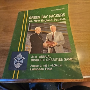 May include: A vintage Green Bay Packers vs. New England Patriots game program. The cover is green with yellow stripes and features two men, one holding a football. Text includes "31st Annual Bishop's Charities Game" and "August 3, 1991 - 6:00 p.m. Lambeau Field."