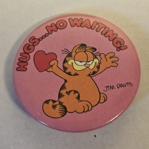 以下が含まれることがあります： ピンク色の丸いバッジ。ガーフィールドという猫が赤いハートを持っています。「HUGS...NO WAITING!」という文字が上部に弧を描いています。アーティストのサイン「JIM DAVIS」が下にあります。