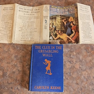 May include: An open book with the title "The Beverly Gray Mystery Stories" and a blue book titled "The Clue in the Crumbling Wall" by Carolyn Keene. The cover of the blue book features an orange silhouette of a person.