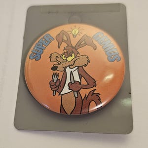 Op de afbeelding: Een ronde oranje button met een cartoon coyote die een vork en mes vasthoudt, met de woorden "SUPER GENIUS" in blauw. De coyote heeft een gloeilamp boven zijn hoofd. De button zit op een grijze achtergrond.