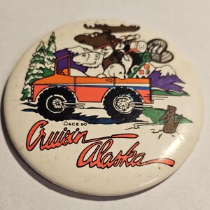 May include: A round, off-white button with a cartoon illustration of an orange truck carrying a moose, bear, and other camping gear. The text "Cruisin' Alaska" is written in red cursive. A pine tree, mountains, and a beaver are also depicted.