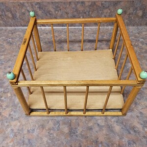 May include: A small, vintage wooden basket with a light brown finish. The basket has a rectangular shape with vertical spindles and a solid base. The corners are adorned with light green knobs. The basket is designed for storage.