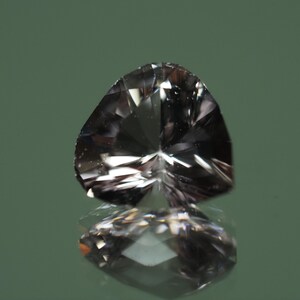 Op de afbeelding: Een gefacetteerde, hartvormige edelsteen met een reflecterend oppervlak. De edelsteen heeft een rookgrijze kleur en ligt op een reflecterend oppervlak, waardoor een spiegelbeeld ontstaat. De achtergrond is een gedempt groen.