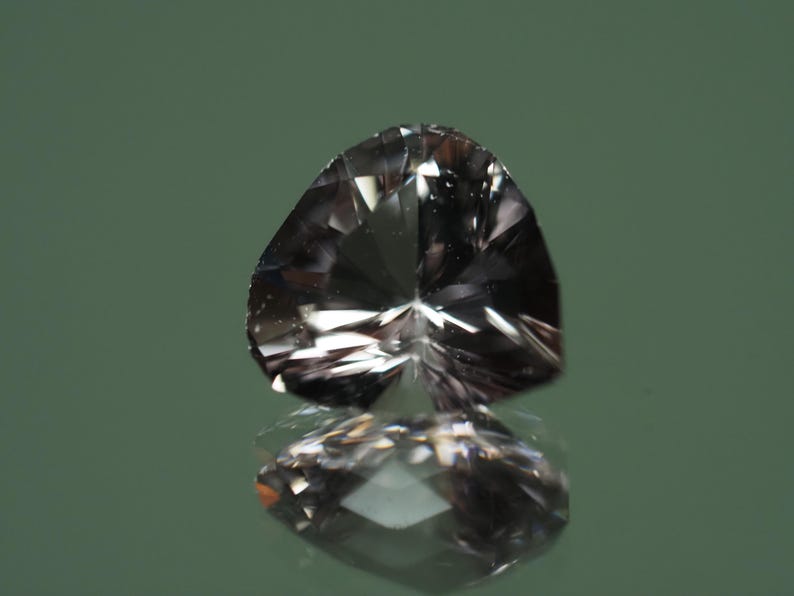Op de afbeelding: Een gefacetteerde, hartvormige edelsteen met een reflecterend oppervlak. De edelsteen heeft een rookgrijze kleur en ligt op een reflecterend oppervlak, waardoor een spiegelbeeld ontstaat. De achtergrond is een gedempt groen.