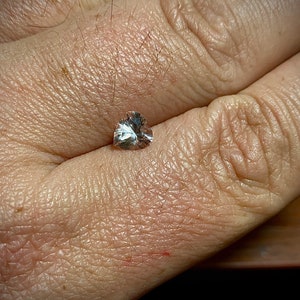 Op de afbeelding: Een kleine, heldere, hartvormige edelsteen met meerdere facetten, rustend op de handpalm van een persoon. De edelsteen reflecteert licht en toont zijn slijpsel en helderheid. De huidskleur is licht, met zichtbare textuur en fijne haren.