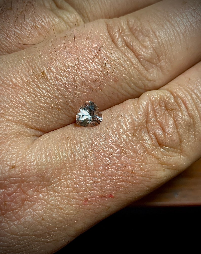 Op de afbeelding: Een kleine, heldere, hartvormige edelsteen met meerdere facetten, rustend op de handpalm van een persoon. De edelsteen reflecteert licht en toont zijn slijpsel en helderheid. De huidskleur is licht, met zichtbare textuur en fijne haren.