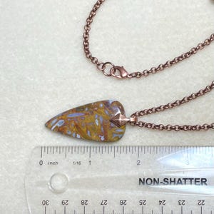 Op de afbeelding: Een koperen ketting met een hanger in de vorm van een pijlpunt van bruine, witte en blauwe agaat. De hanger is ongeveer 5 cm lang. De ketting wordt gemeten op een liniaal met de tekst "NON-SHATTER" erop gedrukt.