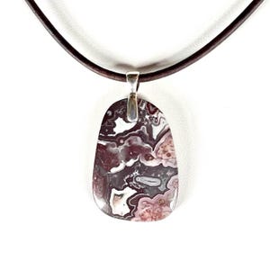 May include: A pendant necklace featuring a polished, oval-shaped stone with a unique pattern of maroon, white, and gray swirls. The stone is suspended from a silver-colored bail and a dark brown leather cord. The necklace is displayed against a white background.