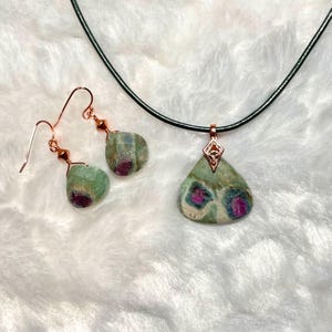 May include: A green and pink gemstone necklace and earrings set with a black leather cord. The pendant and earrings feature a teardrop-shaped stone with pink and green inclusions.