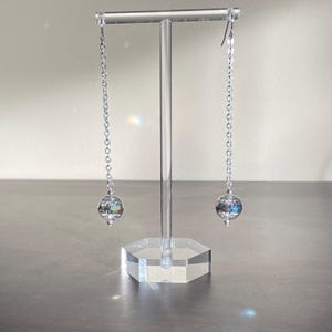 May include: A pair of silver dangle earrings with clear crystal ball charms. The earrings are hanging from a clear acrylic stand.