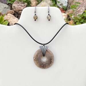 May include: A necklace and earring set featuring a donut-shaped pendant with a brown and cream marbled pattern. The pendant is attached to a black cord and has a silver-colored metal accent. The earrings have matching beads.