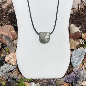 May include: A green and silver stone pendant on a black leather cord necklace. The pendant is a rectangular shape with rounded corners.
