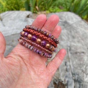 May include: A set of six bracelets featuring a mix of purple and copper colored beads, including a braided leather bracelet.